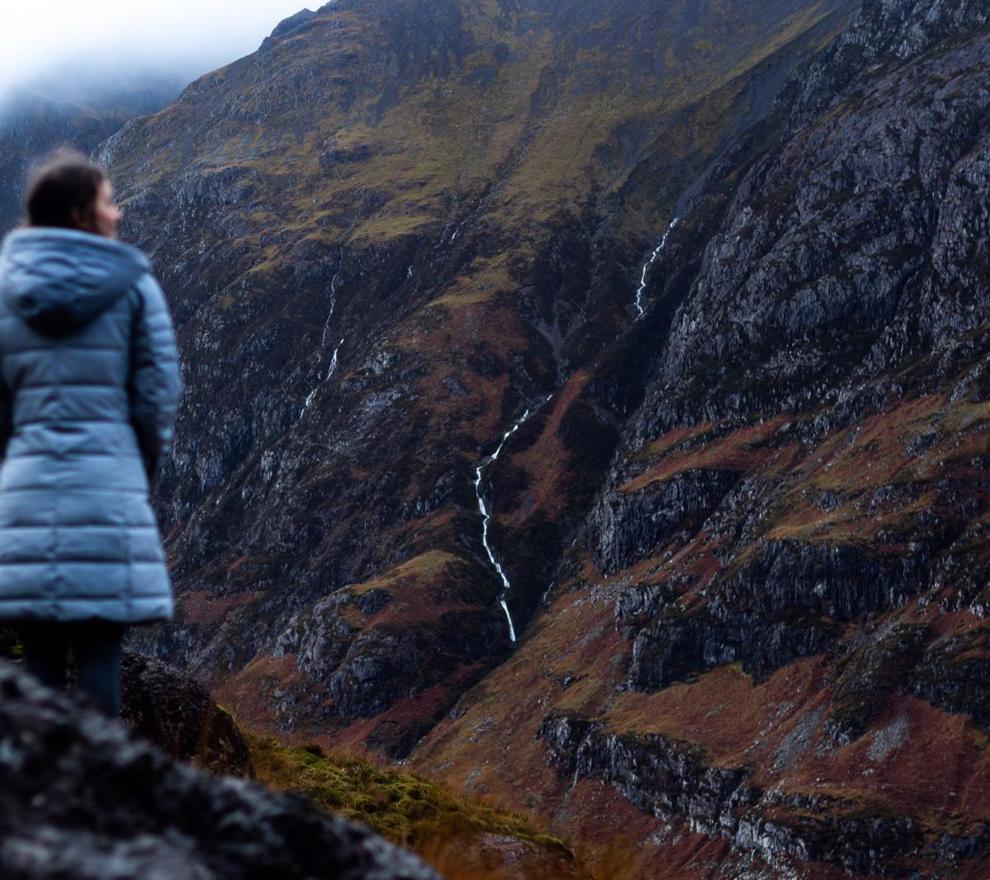 A woman wearing a puffer coat stands overlooking a dramatic, rugged mountain landscape in Glencoe, Scottish Highlands, with thin waterfalls cascading down the slopes. ?? ''