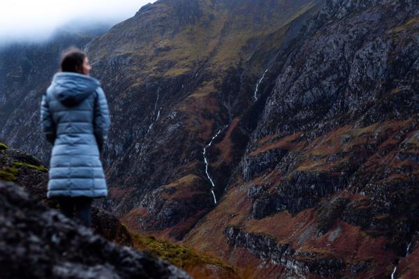 A woman wearing a puffer coat stands overlooking a dramatic, rugged mountain landscape in Glencoe, Scottish Highlands, with thin waterfalls cascading down the slopes. ?? ''