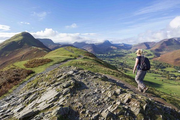 Woman hiking on a rocky hilltop path overlooking rolling green valleys and distant peaks in the Lake District on a clear, sunny day. ?? ''