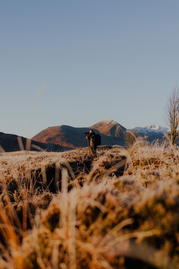 Photographer with a camera on a tripod capturing a frosty morning landscape in Glencoe, Scotland, with snow-dusted mountains in the background. ?? ''