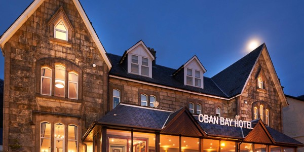 Exterior view of the Oban Bay Hotel, a traditional stone building with a modern glass extension, illuminated at dusk with a clear night sky. ?? ''