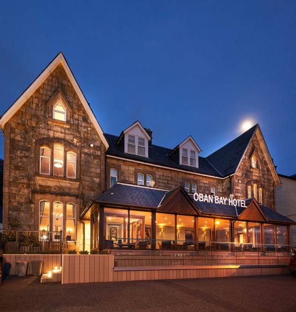 Exterior view of the Oban Bay Hotel, a traditional stone building with a modern glass extension, illuminated at dusk with a clear night sky. ?? ''